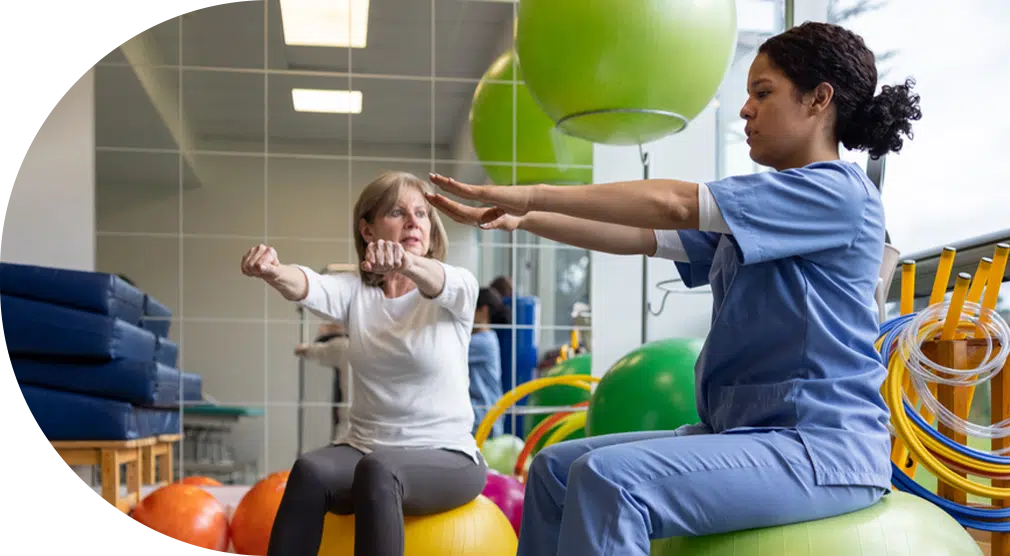 Young woman nurse of the Centers for Advanced Orthopaedics in scrubs sitting on a yoga ball and holding her arms out to instruct her patient, to the left of her, who is doing the same movement.