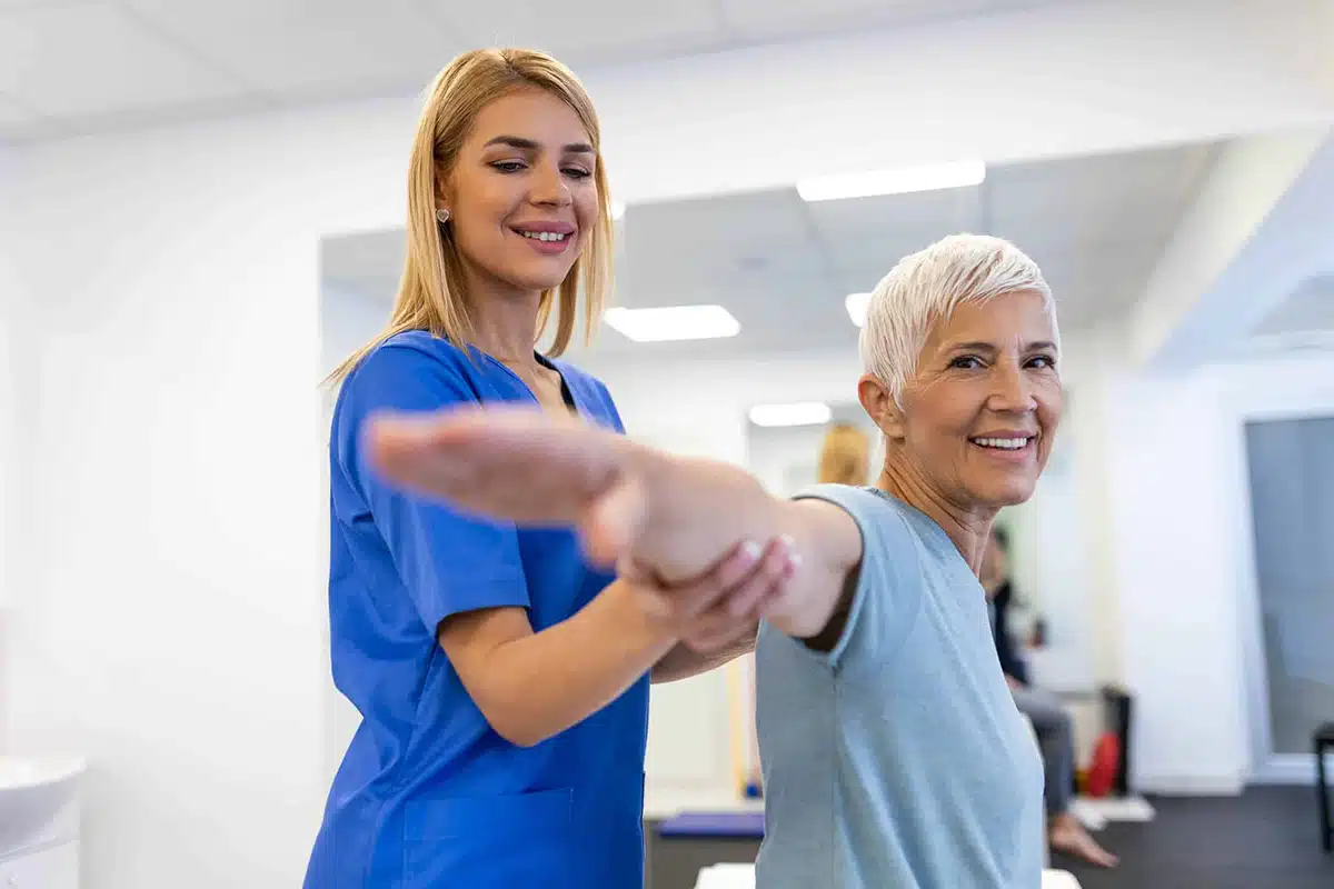 Young woman nurse helping her older woman patient stretch her arms out.