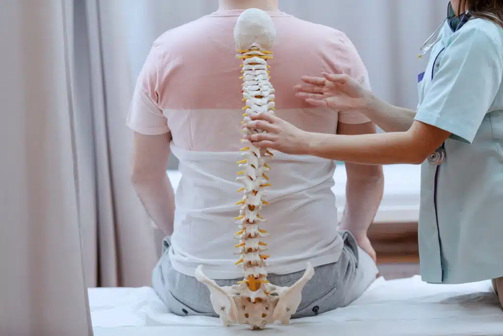 A female orthopedic back specialist holds a spine model against a patient's back who is experiencing back pain at the clinic.