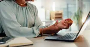Image of a woman suffering from hand and wrist injuries while using her laptop in the office, experiencing pain and discomfort due to carpal tunnel syndrome.